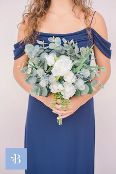 a woman in a blue dress holding a bouquet of white flowers and greenery on her arm