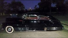 an old black car parked on the side of the road at night with it's lights on