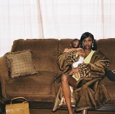 a woman sitting on a couch holding a baby in her arms and wearing a fur coat