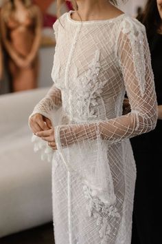 a woman wearing a white dress with sheer sleeves and flowers in her hair standing next to another woman