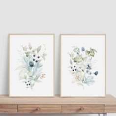 two watercolor flowers are displayed on the wall in front of a wooden table with drawers