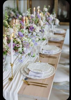 the table is set with white plates and purple flowers on them, along with candles