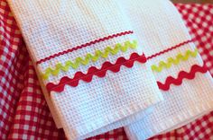 two white towels with red, green and yellow trims on them sitting on a checkered table cloth