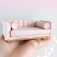 a hand holding a miniature pink couch with pillows on it's sides and hearts flying in the background