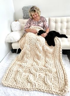 an older woman sitting on a white couch holding a black cat under a knitted blanket