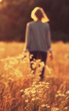 a woman walking through a field with tall grass and wildflowers in the foreground