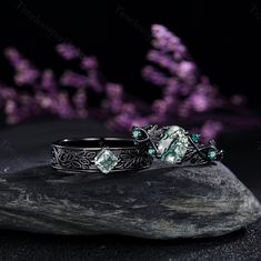 two silver rings with green stones sitting on top of a rock next to purple flowers