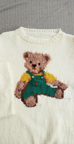 a white sweater with a brown teddy bear on it's chest and green overalls