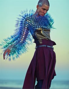 a woman with blue feathers on her head standing near the ocean and wearing purple pants