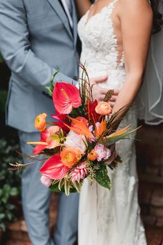 a bride and groom standing next to each other in front of a brick wall holding a colorful bouquet