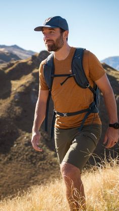 a man with a hat and backpack walking through tall grass in front of mountains on a sunny day