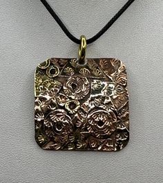 a square pendant with an intricate design on a black cord necklace hanging from a white wall