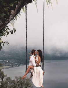 two people are sitting on a swing in front of the water and trees, one person is kissing the other