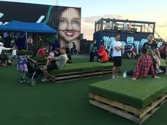 people are sitting and standing around in the grass at an outdoor event with large billboards behind them