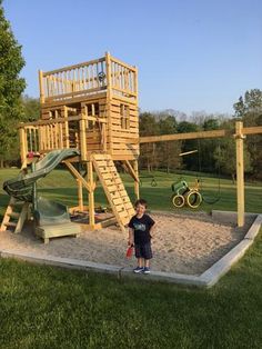 a young boy standing in front of a wooden play structure with slide and climbing frame