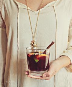 a woman holding a glass filled with liquid and a wooden stick in her hand while wearing a necklace