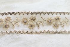 a white lace with gold flowers and beads on it's edge is laying on a piece of fabric