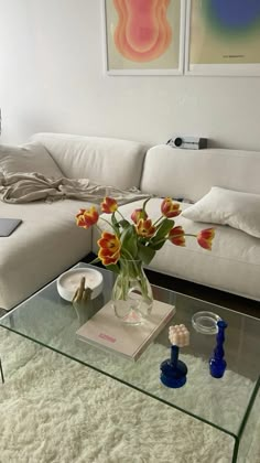 a glass table with flowers on it in front of a white couch and two paintings