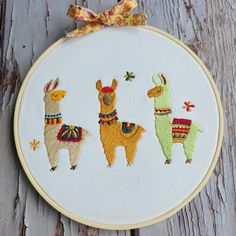 three llamas in different colors and patterns on a white hoop hanging from a wooden surface