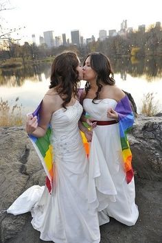 two women in white dresses are kissing each other with rainbow colors on their capes