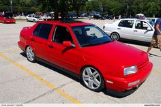 a red car parked in a parking lot next to other cars and people walking by