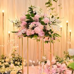 an arrangement of pink flowers and greenery on a gold stand with candles in the background