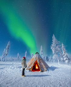 a person standing in front of a small cabin with an aurora light above it and snow covered trees