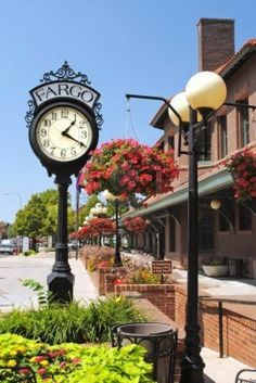 a clock on a pole in front of some flowers and buildings with potted plants