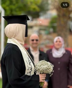 a woman wearing a graduation gown and holding a bouquet of flowers in front of other people