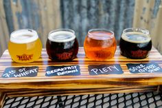 four different types of beer are on a wooden tray with chalk writing and the names of their beers