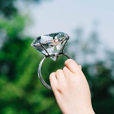 a person holding a ring with a diamond in it's middle and trees in the background