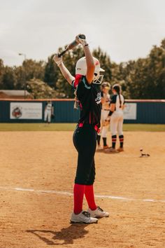 a girl in black and red uniform holding up a baseball bat on a field with other players behind her