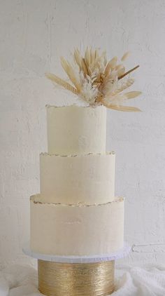 a three tiered white and gold wedding cake with feathers on top, sitting on a table