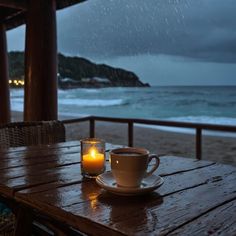 two coffee cups on a wooden table next to the ocean with a candle in front of them