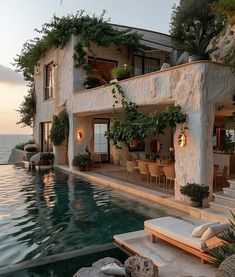 an outdoor pool with chairs and tables next to the water at sunset or dawn in front of a house