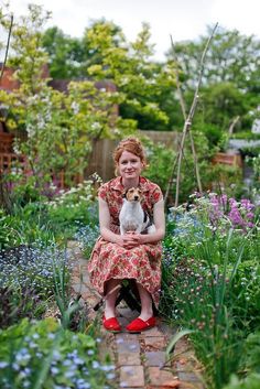 a woman sitting in the middle of a garden with her small dog on her lap