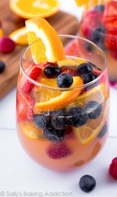 a glass filled with fruit and ice on top of a white table next to sliced oranges, raspberries and blueberries