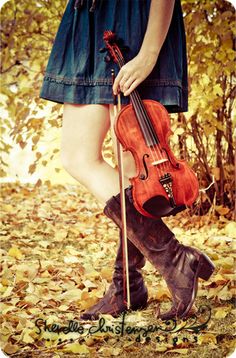 a woman in boots holding a violin on top of leaf covered ground with trees behind her