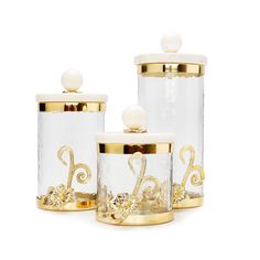 three glass containers with gold accents and pearls on the lids, one has a monogrammed letter