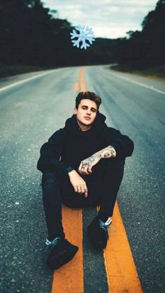 a young man sitting on the side of an empty road with his hands in his pockets