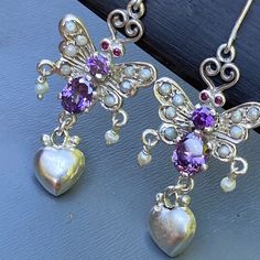 Very Well handmade ~ Victorian inspired earrings with absolutely fabulous details ~ Earrings are newly designed and are inspired by Victorian earrings using higher grade silver ,faceted Amethyst stones , paved seed pearls and garnet eyes with puffed heart dangles at bottom ~ Unmarked sterling grade silver . Measurements ~ Earrings are 1 3/4 inches in total drop length x max width of 3/4 inches . Combine weight of earrings is 5 grams on my weighing scale. Condition : As earrings are had made plea Ornate Purple Sterling Silver Earrings, Egyptian Revival Jewelry, Butterfly Dangle Earrings, Victorian Earrings, Amethyst Stones, Bali Silver, Puffed Heart, Weighing Scale, Absolutely Fabulous