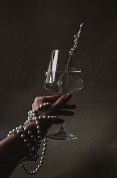 a person holding a glass with pearls and a chain attached to it, in front of a dark background