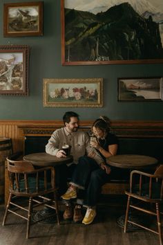 a man and woman sitting at a table in a room with paintings on the wall