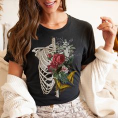 Welcome to Guava Zone! This beautiful ribcage t-shirt is the perfect tee for your flower loving friend. This floral inspired graphic tee is the best way to show off your boho style and love for flowers, with a cool twist on our usual skeleton design! Our designs are printed on superior soft cotton tees. This classic unisex jersey short sleeve t-shirts have ribbed elastane collars and reinforcing tape on neck to hold the garment's shape for longer. This style is unisex and it runs true to size. R Black Bohemian Tops For Halloween, Black Bohemian Top For Halloween, Black Bohemian Top For Alternative Fashion, Ribcage Shirt, Botanical Anatomy, Floral Ribcage, Anatomy Shirts, Human Bones, Skeleton Design