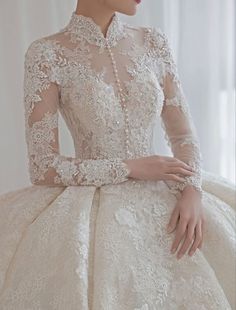 a woman wearing a wedding dress with long sleeves and lace on the top, sitting in front of a window