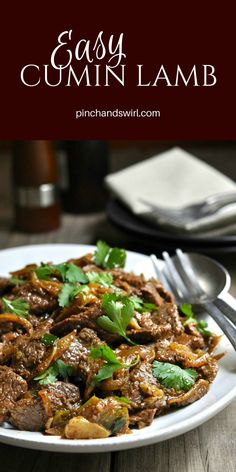 a white plate topped with beef and cilantro next to silverware on a wooden table