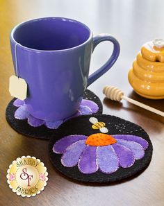 two coasters with purple flowers on them next to a honey comb and a mug