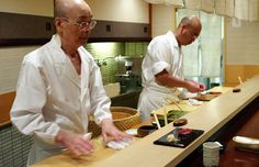 two men preparing food in a kitchen on top of a wooden counter next to each other