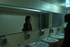 a woman standing in front of a mirror next to two sinks and urinals