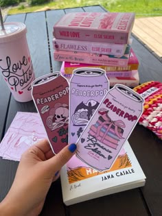 a person is holding up four pink bookmarks on a picnic table with coffee and books